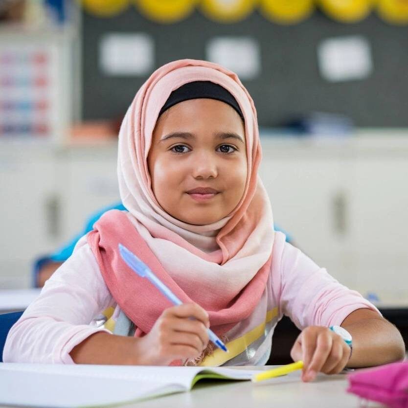 A girl in a pink hijab is writing at her desk.